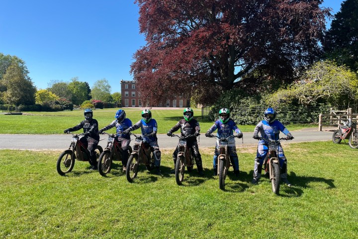 Six people on dirt bikes lined up on grass with trees and a building in the background.