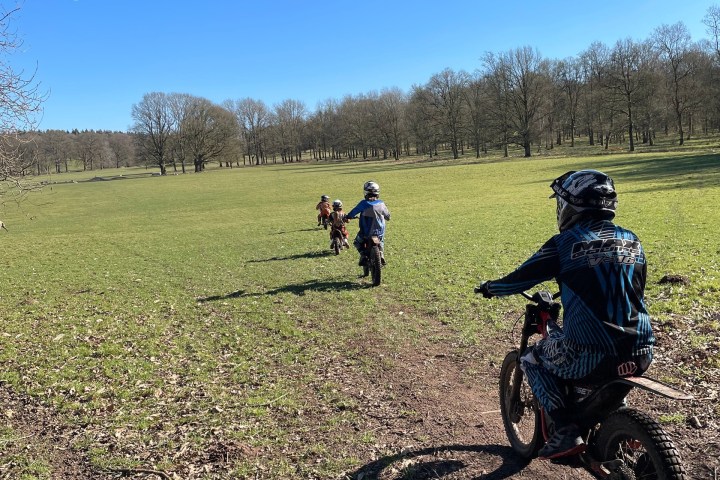 Children ride dirt bikes on a grassy trail in open field under clear blue sky.