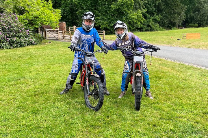 Two people in motocross gear standing with bikes on grassy field.